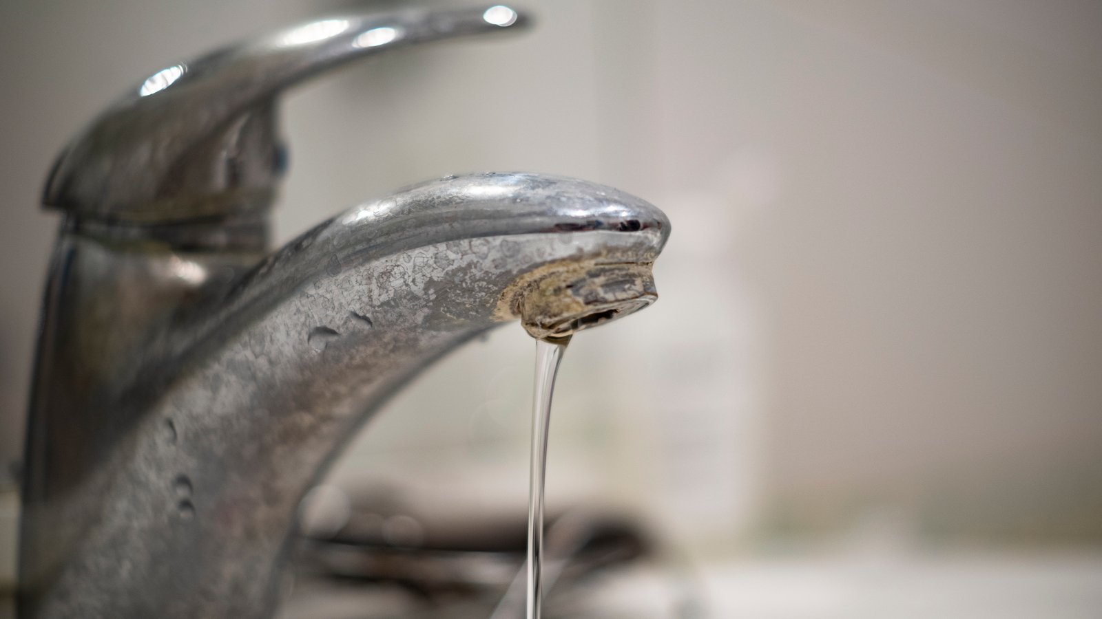 Close-up of a chrome faucet spout with hard water mineral buildup crusted around the opening and a thin stream of water running out